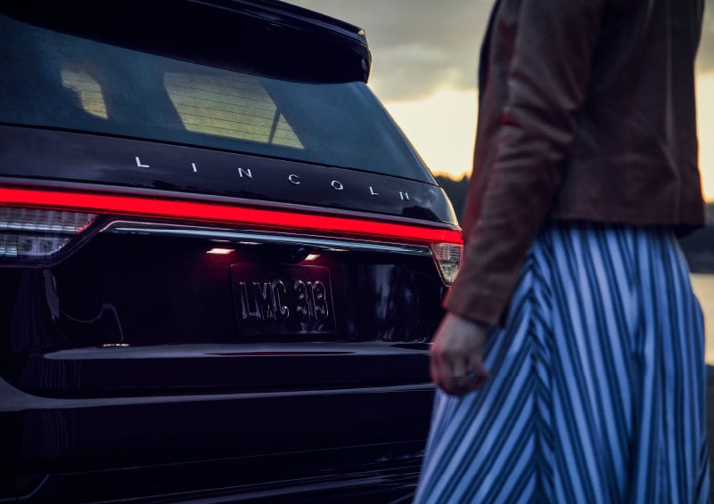 A person is shown near the rear of a 2024 Lincoln Aviator® SUV as the Lincoln Embrace illuminates the rear lights | Brinson Lincoln of Athens in Athens TX